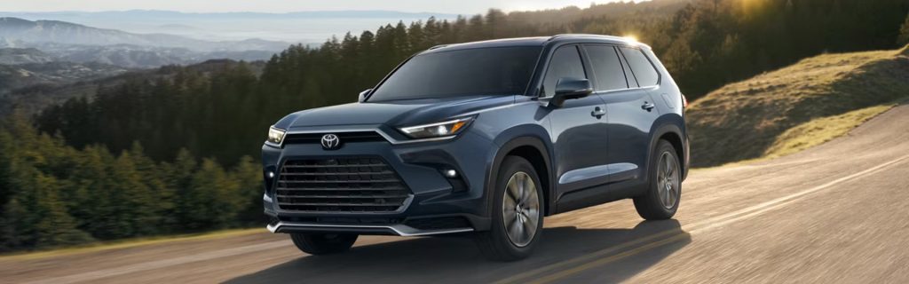 A blue Toyota Grand Highlander driving on a scenic road surrounded by green trees and hills.